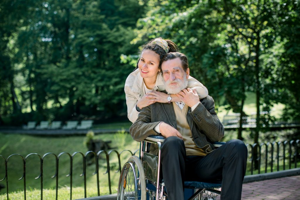 Happy bearded senior man in wheelchair with charming young girl supporting disabled grandparent on